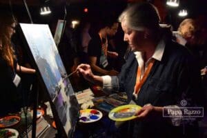 An artist holding a paint palette and a paint brush painting an outdoor landscape on canvas