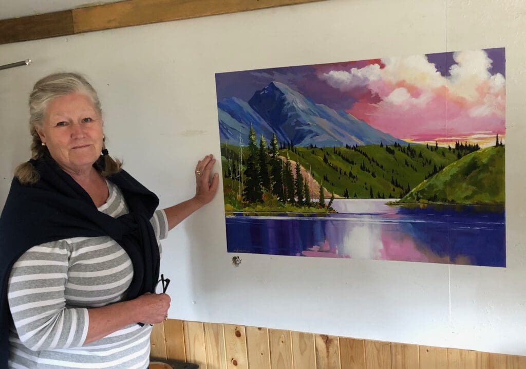 An artist posing beside her painting of a mountain lake at dusk with a pretty pink and purple sky