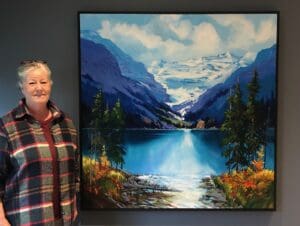An artist smiling standing beside her painting of a mountain landscape